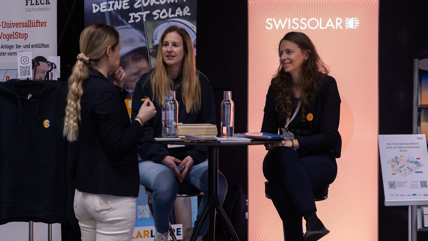 Drei Frauen unterhalten sich an einem Stand mit orangenem Hintergrund | © Swissolar / Sarah Bollmann